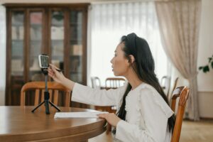 A young woman in white blouse at a table, using a cellphone stand for online learning.