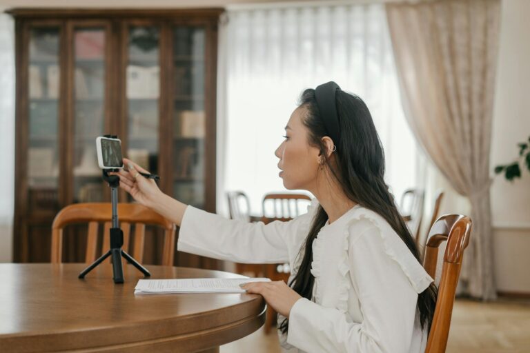 A young woman in white blouse at a table, using a cellphone stand for online learning.